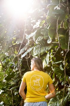 a woman wearing a yellow shirt and blue shorts standing in front of green plants with the sun shining on her back