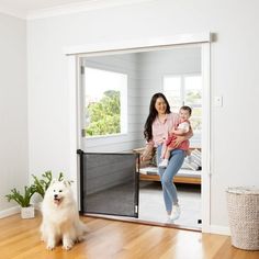 a woman holding a baby standing in front of an open door with a dog on the floor