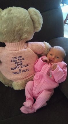 a teddy bear sitting next to a baby wearing a pink sweater with words on it