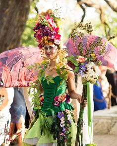 a woman dressed in green holding an umbrella and flowers on her head while walking down the street