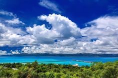 the blue water is surrounded by green trees and white clouds in the sky above it