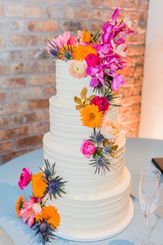 a white wedding cake with flowers on top and wine glasses next to it, in front of a brick wall