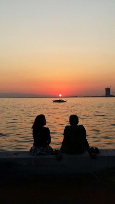 two people sitting on the edge of a pier watching the sun go down over the water