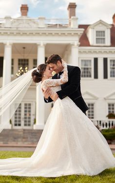 a bride and groom kissing in front of a large white house with a veil blowing in the wind