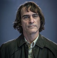 a man with long hair wearing a green shirt and jacket looking at the camera while standing in front of a dark background