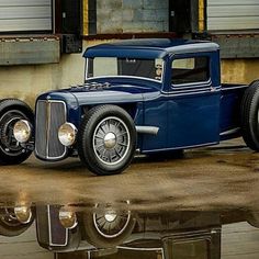 an old blue truck is parked in front of a building and reflecting in the water