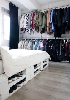 a bed sitting under a window next to a shelf filled with shoes and clothes on hangers