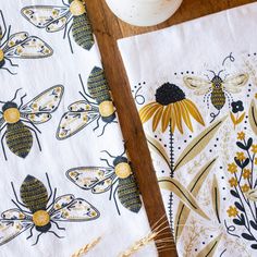 two napkins with bees on them sitting next to a white vase and some yellow flowers