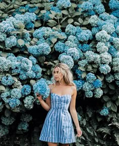 a woman in a blue and white striped dress standing next to a bush full of blue flowers