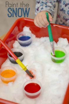 a toddler is painting the snow with paints and brushes in a red tray filled with white foam