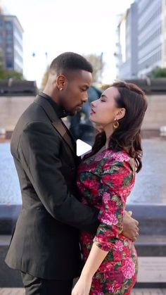 a man and woman standing next to each other in front of a fountain with their arms around each other