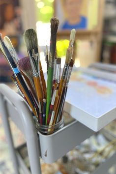 a cup filled with lots of paint brushes on top of a white table next to a painting easel