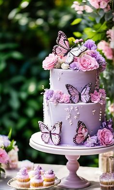 a three tiered purple cake with pink flowers and butterflies on the top is surrounded by cupcakes