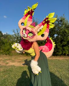 a woman in a green dress is holding a pink dragon mask on her head while standing in the grass