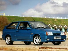 a blue car parked in front of a green hill and white picket fenced area