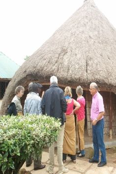 several people standing in front of a thatched roof hut with plants and shrubs around it