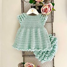 a crocheted baby dress and blooming flowers are hanging on a wire rack