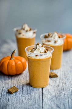 three cups filled with dessert sitting on top of a wooden table next to pumpkins