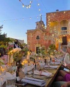 people are sitting at a long table in front of an old building with lights strung from it