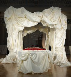 an elaborately decorated stage with white drapes and red roses on the top table