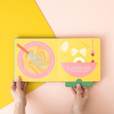 two hands holding an open children's book on top of a yellow and pink background