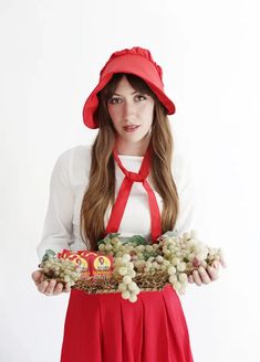 a woman wearing a red hat and holding grapes in her hands while standing against a white background