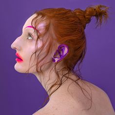 a woman with red hair and ear piercings looking off to the side in front of a purple background