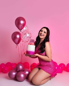 a woman kneeling down with a cake and balloons in front of her on a pink background