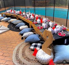 a long table set up with pillows and place settings for an outdoor party next to a swimming pool