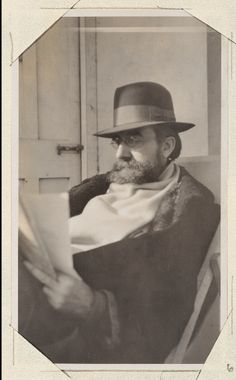 an old black and white photo of a man with a hat on reading a paper