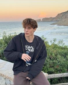 a young man sitting on top of a wooden bench next to the ocean