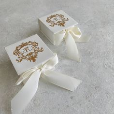 two white boxes with gold designs on them and ribbon tied around the edges, sitting on a gray surface