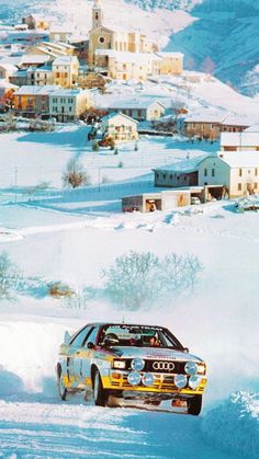 a car driving down a snow covered road in front of houses and buildings on the side of a hill