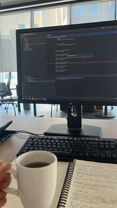 a person holding a cup of coffee in front of a computer monitor and keyboard on a desk