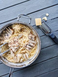 a pan filled with chicken and noodles on top of a wooden table next to a cheese grater