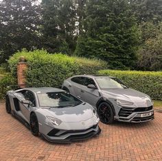 two grey sports cars parked next to each other on a brick driveway in front of bushes