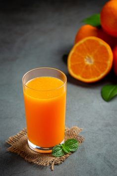 an orange juice in a glass next to some oranges on a gray tablecloth