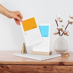 a desk with a calendar on it and a bell in the foreground, next to a vase with flowers