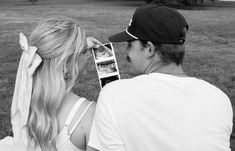 a man and woman sitting on the grass looking at an image in their cell phone