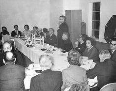 a group of people sitting around a table in front of a man giving a speech