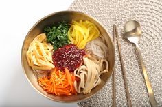 a bowl filled with noodles, carrots and greens next to two spoons on top of a table