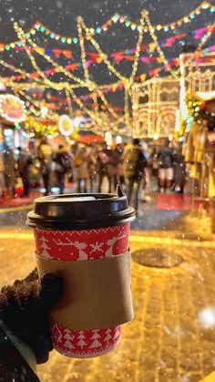 a cup of coffee is held up in front of a christmas scene with lights and people