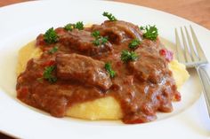 a white plate topped with meat and gravy on top of grits next to a fork