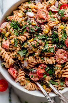 a white bowl filled with pasta salad on top of a marble table next to tomatoes and cilantro