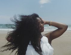 a woman standing on top of a beach next to the ocean with her hair blowing in the wind