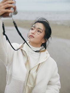 a woman taking a photo with her cell phone on the beach while wearing a white jacket