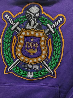 The purple sweatshirt features a large, high-quality chenille patch of the Omega Psi Phi fraternity emblem prominently displayed on the chest. The chenille patch showcases the fraternity's traditional gold and royal purple colors, adding a textured, bold look to the sweatshirt. The patch includes the iconic Omega symbol, surrounded by a Greek-style shield, crossed swords, and other traditional elements representing Omega Psi Phi's legacy of brotherhood, scholarship, and service. The soft, rich purple fabric of the sweatshirt complements the chenille patch, creating a striking and comfortable garment perfect for casual wear or fraternity events. Omega Symbol, Chenille Sweatshirt, Omega Psi Phi Fraternity, Royal Purple Color, Crossed Swords, Chenille Patch, Omega Psi Phi, Purple Sweatshirt, Rich Purple