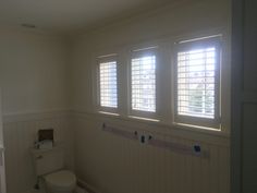 a white toilet sitting next to a window in a bathroom under two windows with blinds on them