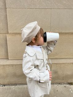 a little boy in a trench coat and hat drinking from a cup while standing next to a wall