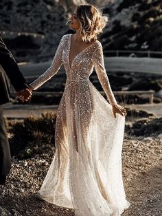 a bride and groom holding hands in front of the mountains at their wedding day, wearing long sleeved gowns with sequin detailing